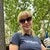 Woman in sunglasses smiling outdoors, wearing a blue shirt with trees in the background.